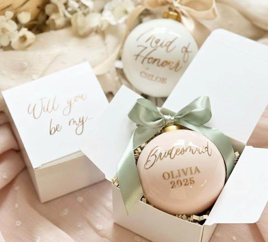 A pale pink ornament with Bridesmaid Olivia 2025 in gold script rests in a gift box with a green bow. Another ornament says Maid of Honour Chloe. A card reads Will you be my... on soft pink fabric.