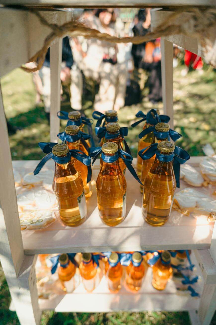 Glass bottles filled with a golden liquid, each topped with a gold cap and tied with a blue ribbon, arranged neatly on white wooden shelves outdoors. Blurred people are visible in the background.