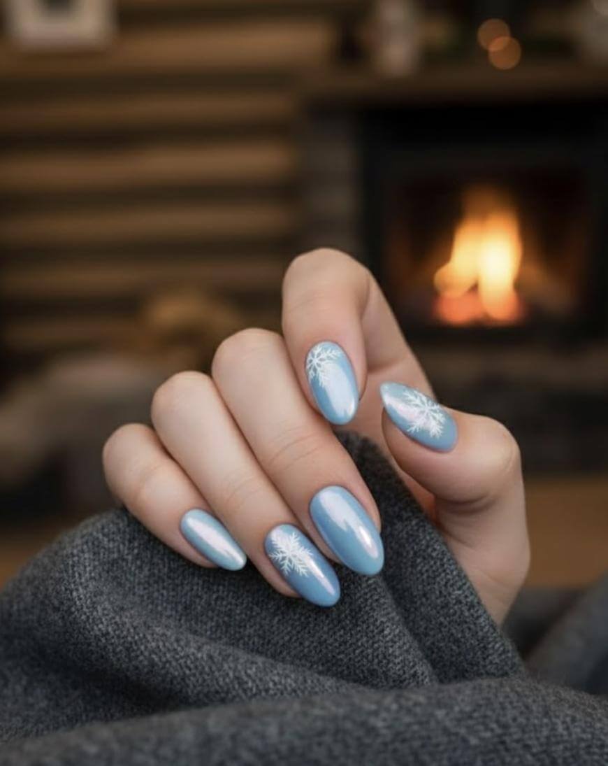 A hand with light blue, almond-shaped nails featuring white snowflake designs is wrapped in a gray blanket. A cozy fireplace with a soft, warm glow is blurred in the background.