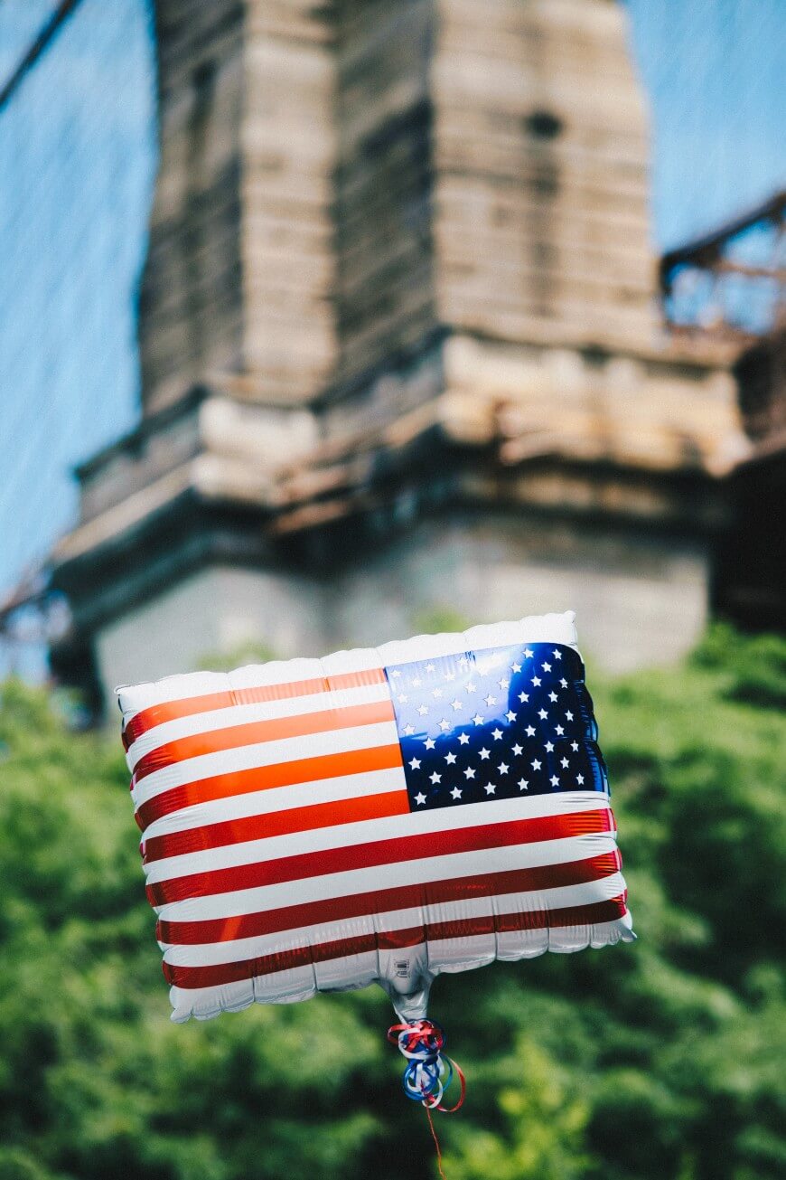 A square balloon decorated with the American flag floats outdoors, with the blurred background of a large stone bridge and green trees.
