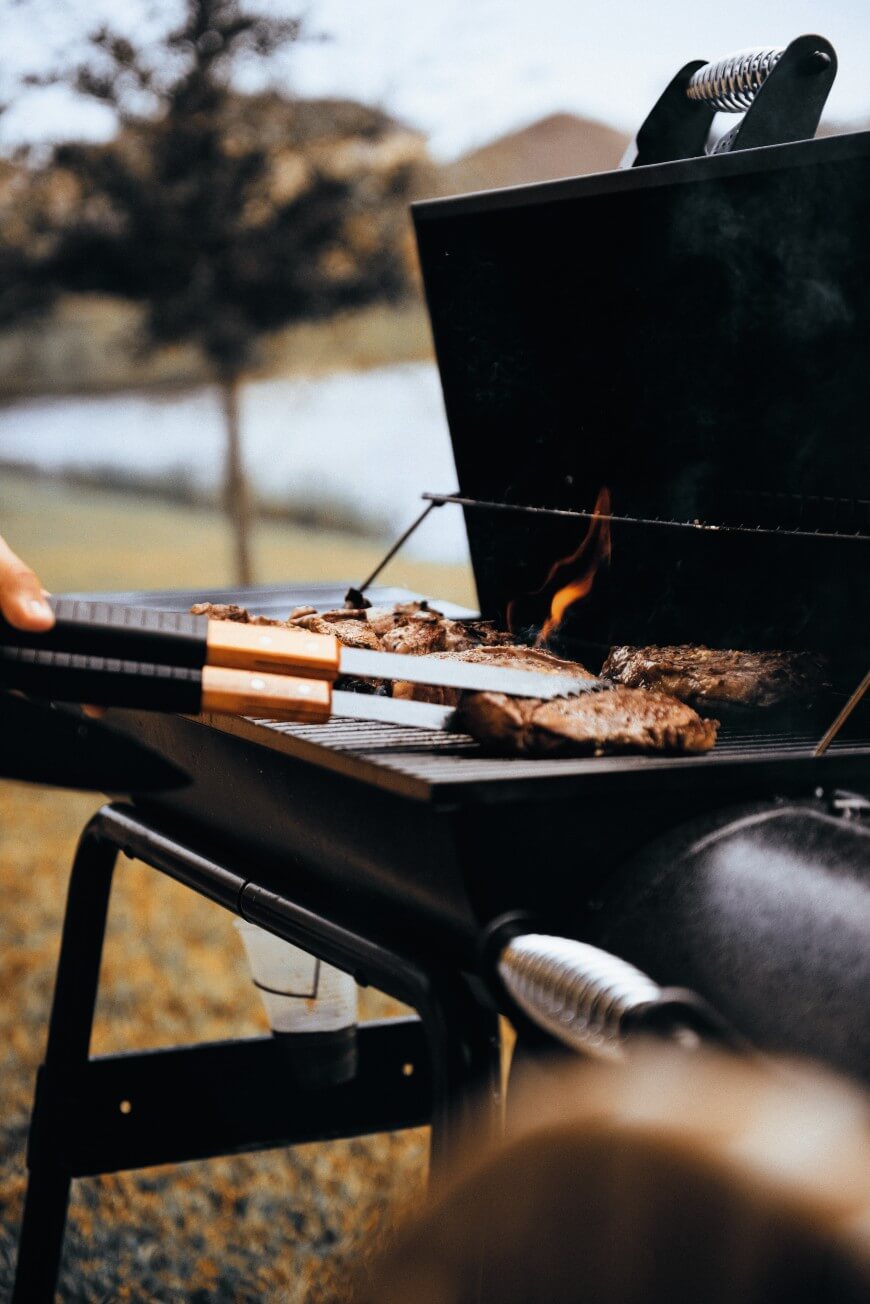 A close-up of a person grilling steaks on an outdoor barbecue grill, with flames visible and a scenic, blurred background of trees and grass.