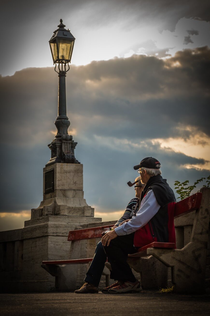 Two elderly people sit on a red bench near a stone railing, with a vintage streetlamp beside them and dramatic, cloudy skies at sunset in the background.