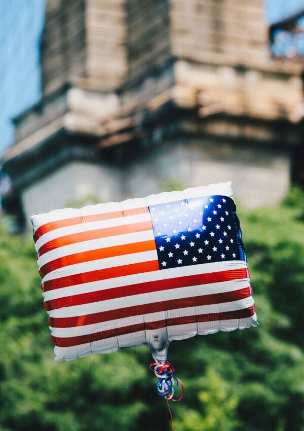 A square balloon decorated with the American flag floats outdoors, with a blurred stone structure and green trees in the background.