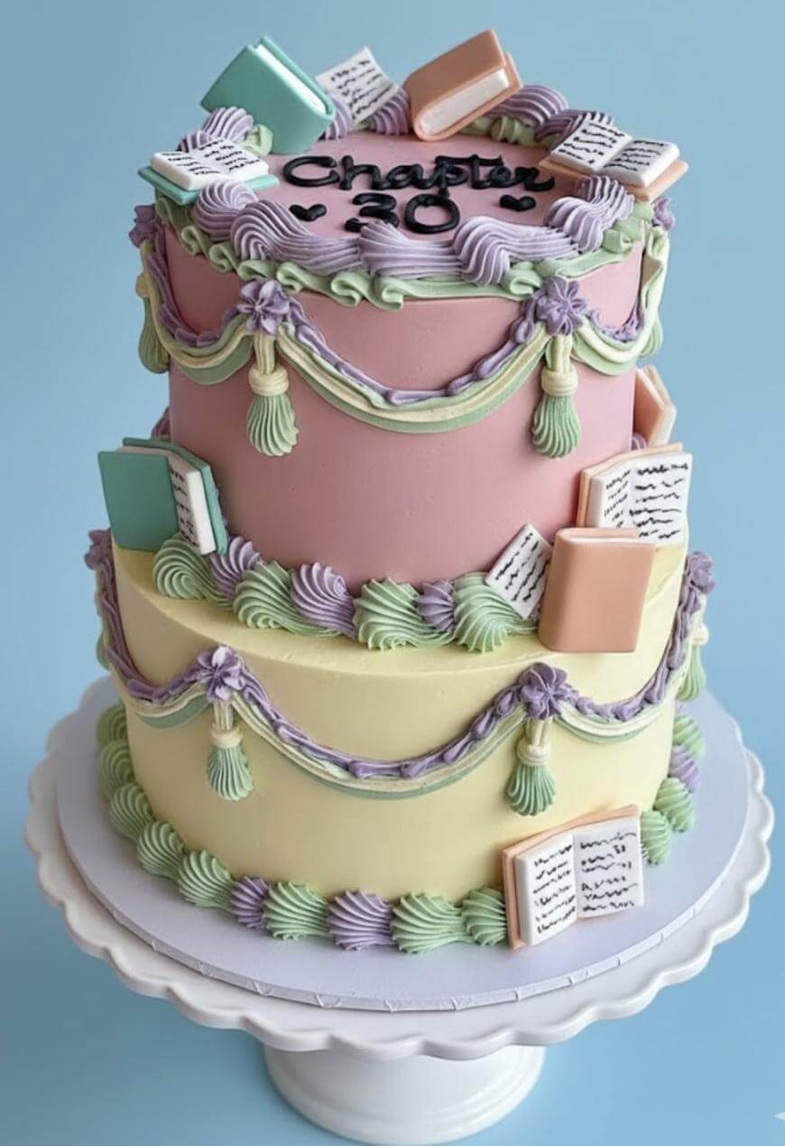 A two-tier pastel cake decorated with icing garlands and fondant books, some open and some closed. The top tier is inscribed with “Chapter 30” in black icing. The cake sits on a white cake stand.