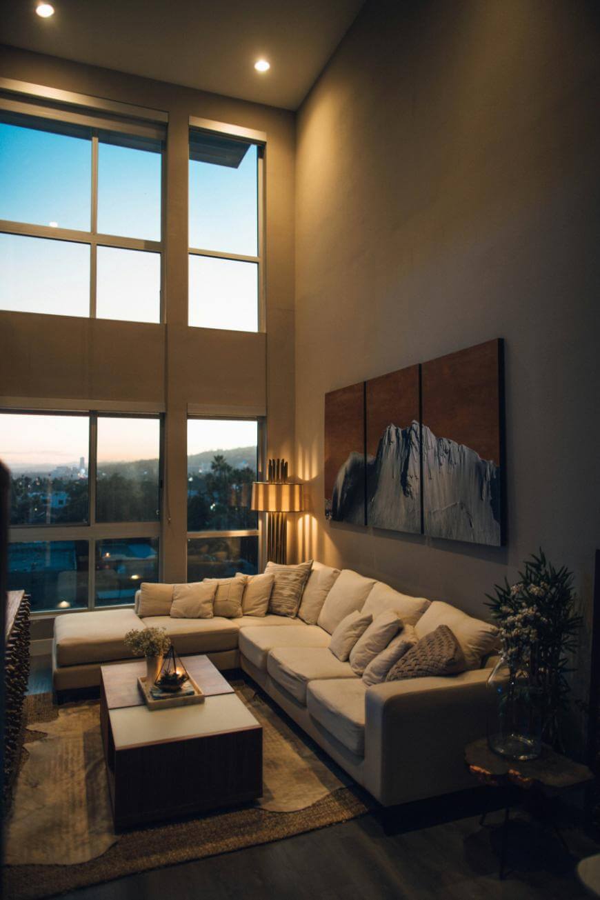 A modern living room with tall windows, a large cream sectional sofa, a wooden coffee table, wall art, indoor plants, and warm ambient lighting during sunset.