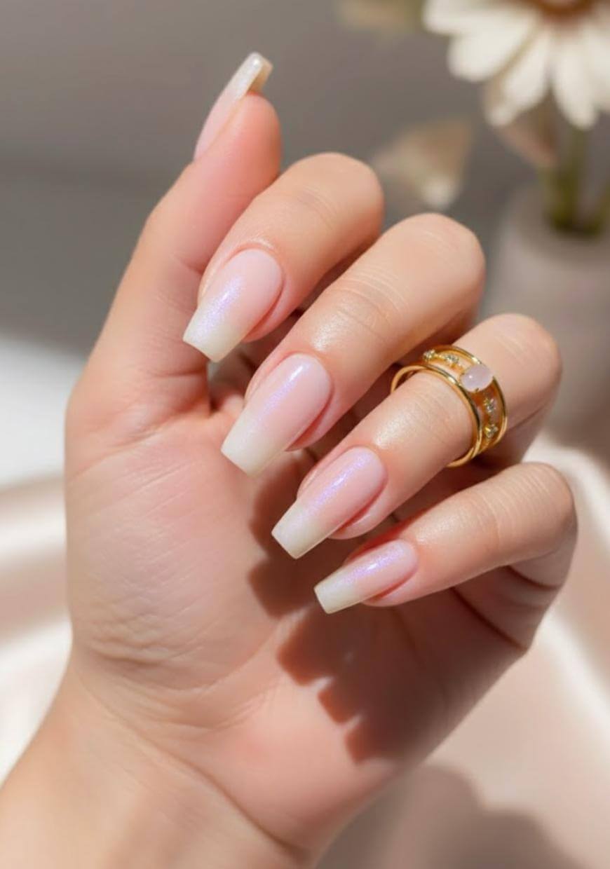 A hand with long, square-shaped, light pink acrylic nails and a gold ring adorned with small stars. The background is softly blurred, with hints of flowers visible.