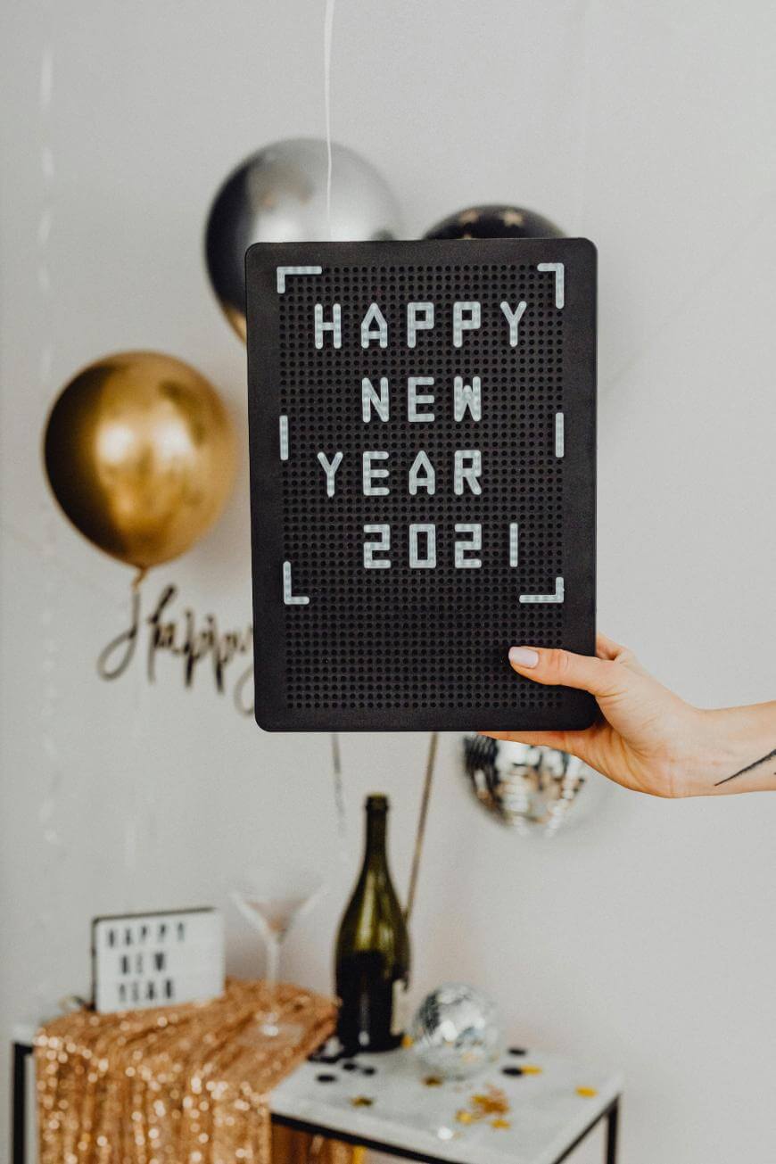 A hand holds a black letter board that reads Happy New Year 2021, with gold, black, and silver balloons and festive decorations in the background.