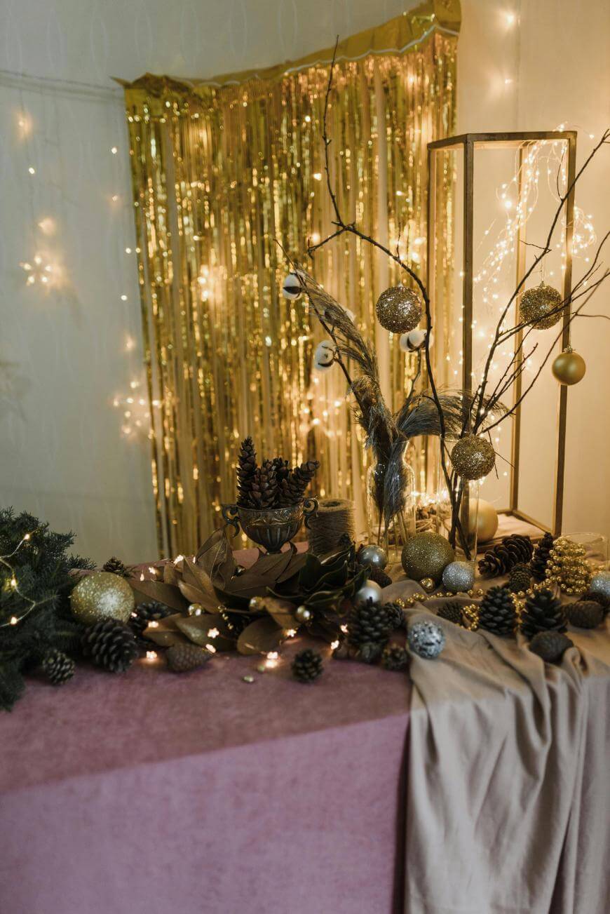 A festive table display with pinecones, golden ornaments, and fairy lights, set in front of a gold tinsel backdrop and decorative branches. The table is covered with a pink and beige cloth.