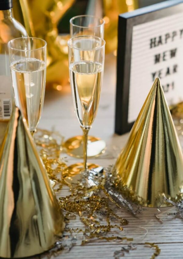 Two glasses of champagne, gold party hats, gold confetti, and a bottle are on a table. In the background, a sign reads Happy New Year 2021. The scene suggests a festive New Year’s celebration.