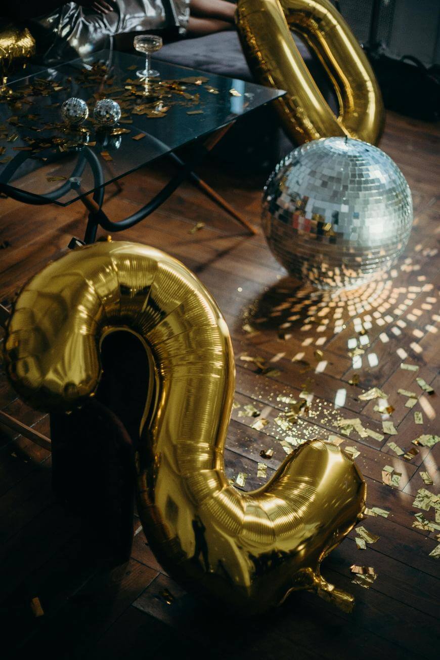 A room with a wooden floor covered in gold confetti, a large gold foil balloon shaped like the number two, a silver disco ball reflecting light, and a table with drinks and shiny decorations.