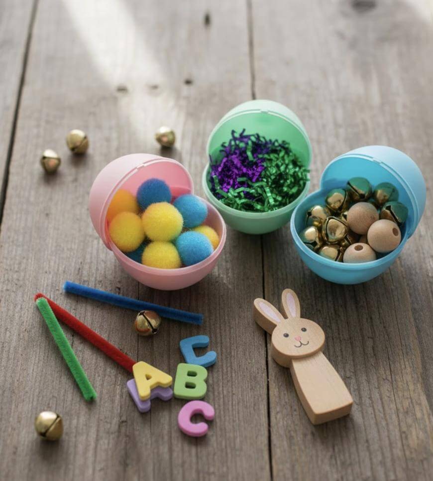 Three plastic Easter eggs filled with colorful pom-poms, shredded paper, and beads sit on a wooden surface with pipe cleaners, small bells, foam alphabet letters, and a wooden bunny nearby.