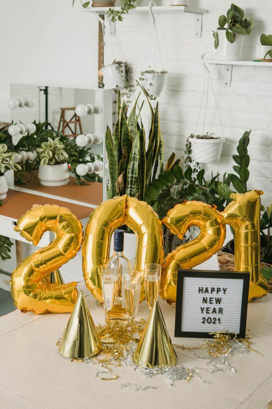 Gold balloons spelling 2021 are surrounded by green houseplants, party hats, two wine glasses, a wine bottle, and a sign reading Happy New Year 2021 on a decorated table in a bright room.