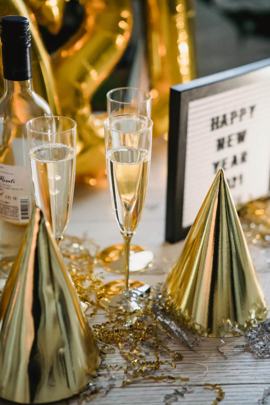 Two glasses of champagne, a wine bottle, gold party hats, and festive decorations sit on a table next to a sign that reads Happy New Year 2021. Gold balloons are blurred in the background.