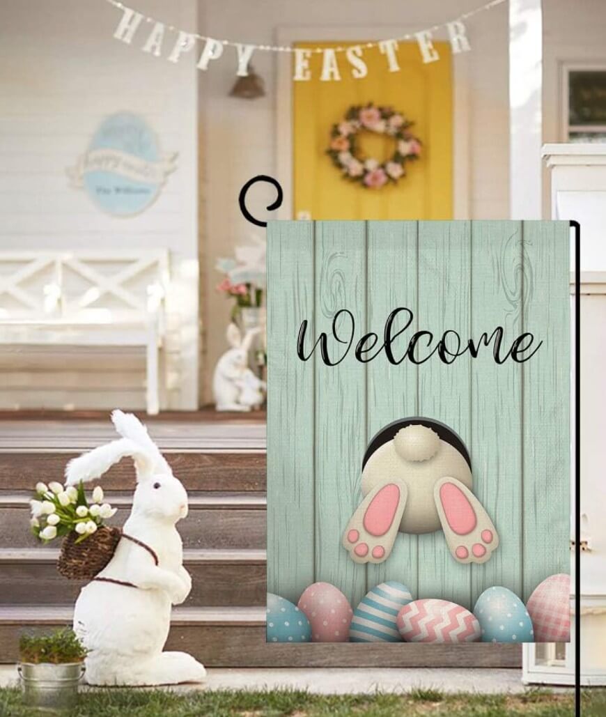 A decorative Easter flag with a bunny’s backside and colorful eggs says “Welcome.” In the background, a porch features a large bunny decoration, a wreath, and a “Happy Easter” banner.