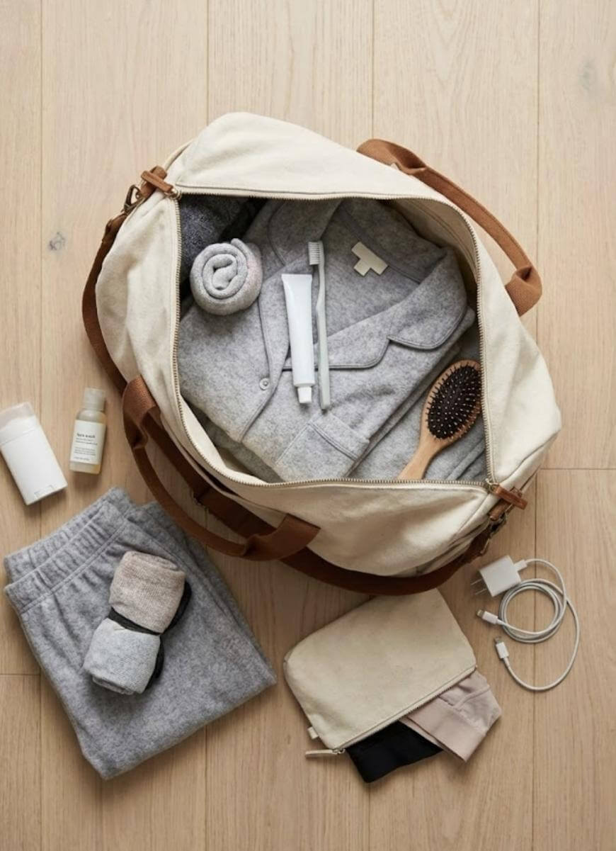 A beige duffel bag on a wooden floor, packed with neatly folded gray clothes, a rolled towel, toothbrush, toothpaste, wooden hairbrush, and travel-size toiletries. Next to the bag are socks, sweatpants, a pouch, and charging cables.