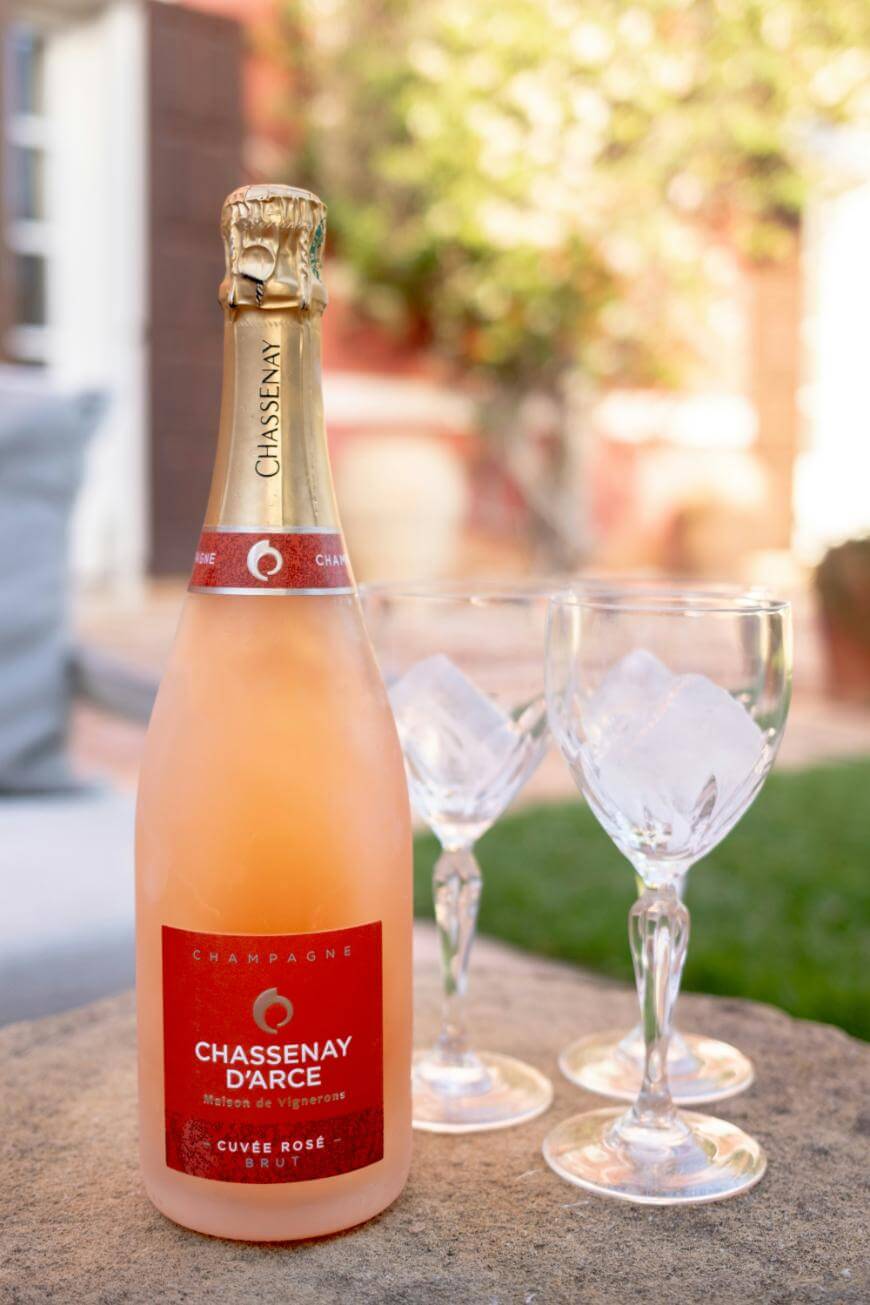 A bottle of Chassenay d’Arce Cuvée Rosé Brut champagne sits on a stone surface outdoors, next to two empty wine glasses with ice, with a blurred garden background.