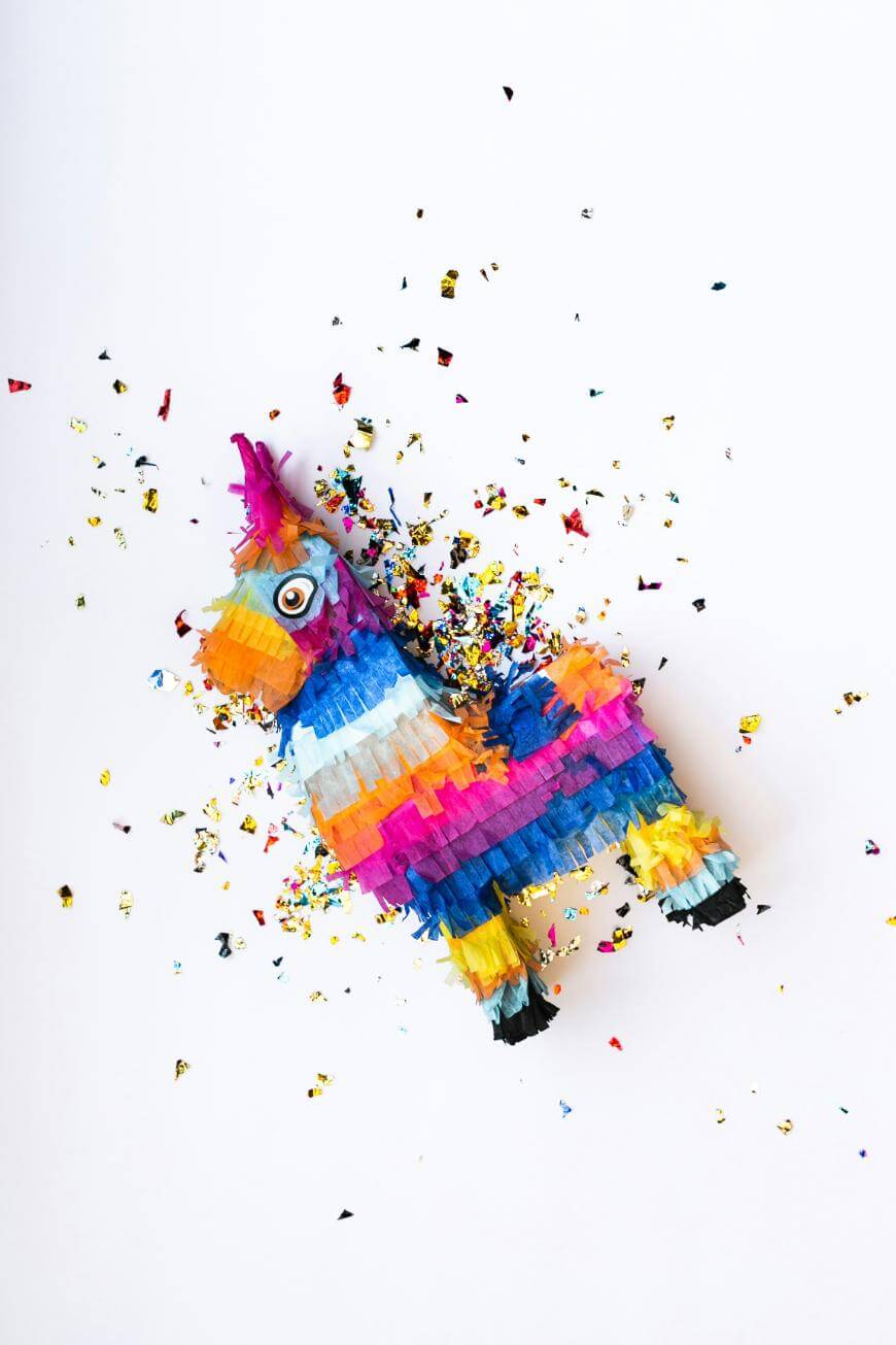 A colorful unicorn piñata with a googly eye lies on a white background, surrounded by scattered multicolored confetti.