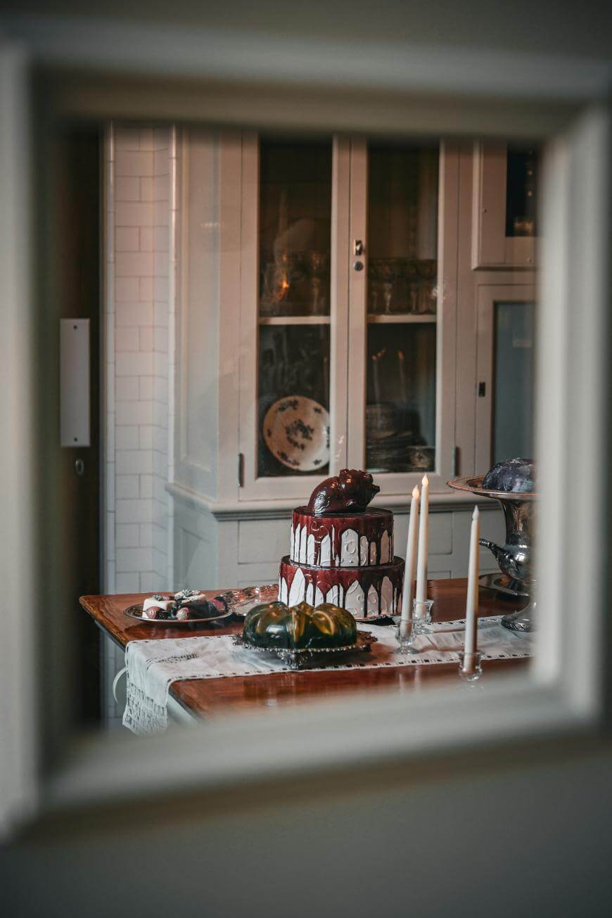 A dark chocolate cake with red dripping icing and a heart decoration sits on a wooden table with candles and other treats, viewed through a window into a vintage-style kitchen.