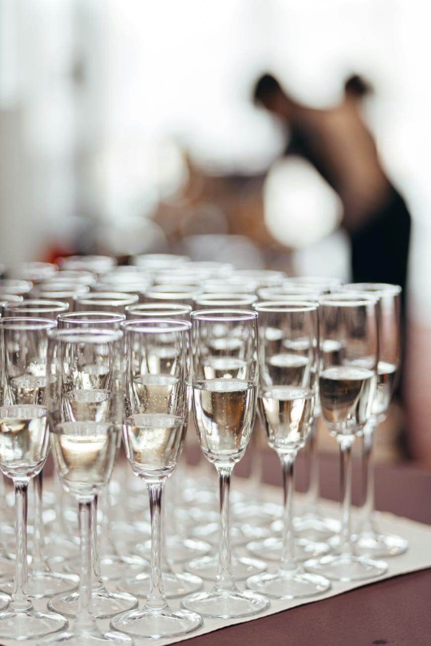Rows of filled champagne glasses are arranged neatly on a table, with a blurry figure in the background, creating a festive and elegant atmosphere.