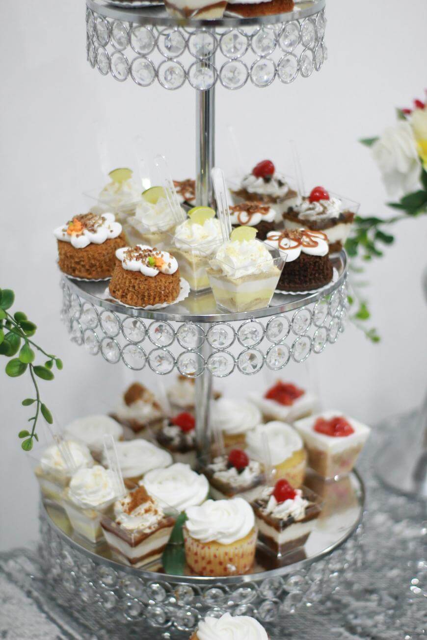 A three-tiered crystal stand displays assorted mini desserts, including cupcakes, parfaits, and cakes, each topped with cream, fruit, or nuts. Greenery and flowers decorate the elegant setup.