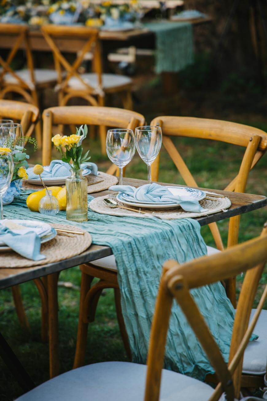 A rustic outdoor table is set with wooden chairs, blue napkins, glassware, small yellow flowers in vases, lemons, and a textured blue table runner. The setting suggests a festive, elegant garden party or event.