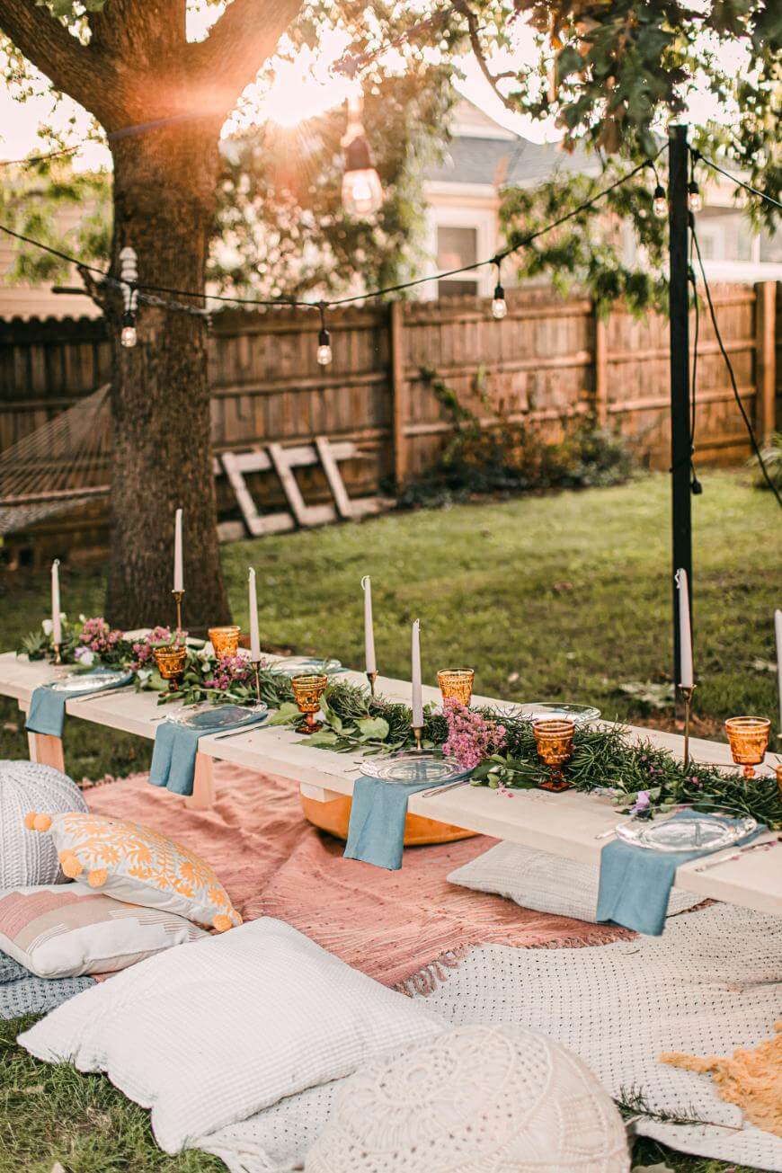 A low outdoor table set for a meal in a backyard, decorated with flowers, candles, and amber glasses. Cushions and pillows are arranged for seating on a patterned rug under hanging string lights.