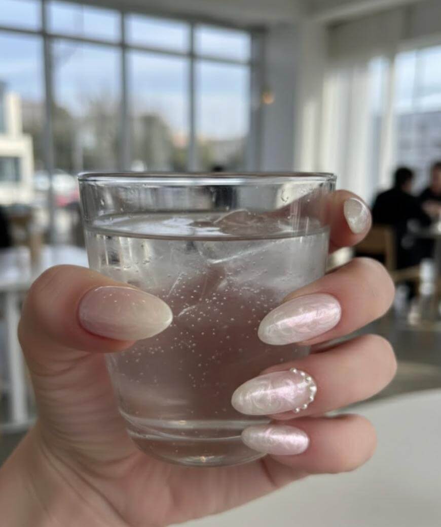 A hand with long, pastel pink and white marble nail art holds a clear glass of sparkling water in a bright, modern café with large windows in the background.