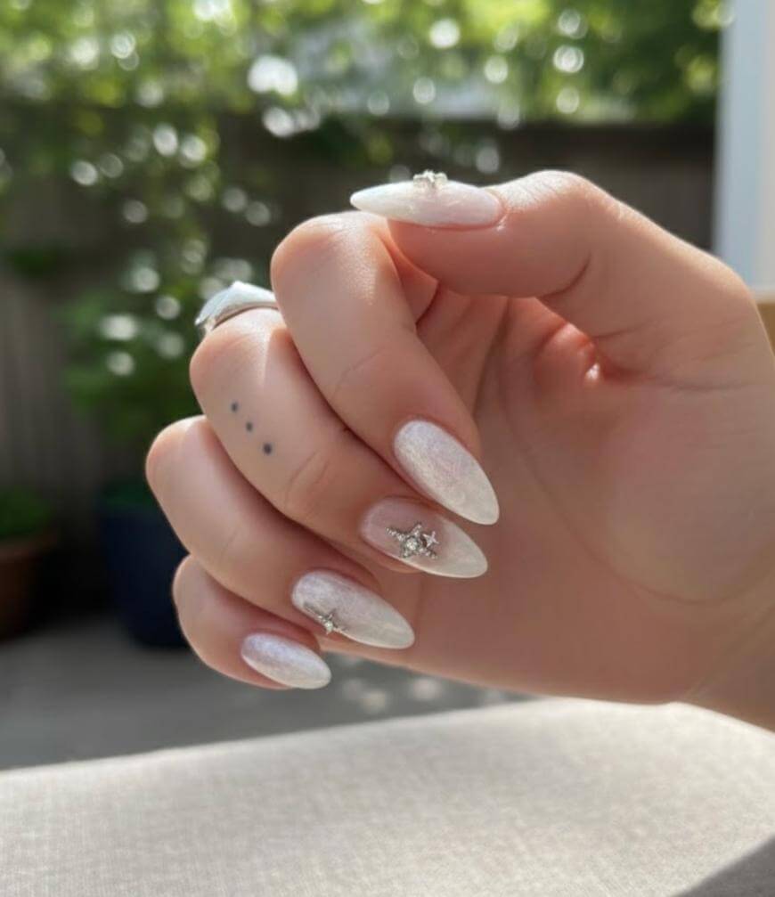 A hand with long, almond-shaped nails painted in shimmery white polish, some adorned with small silver star and moon charms. The hand displays three small dot tattoos on the finger. The background is softly blurred greenery.
