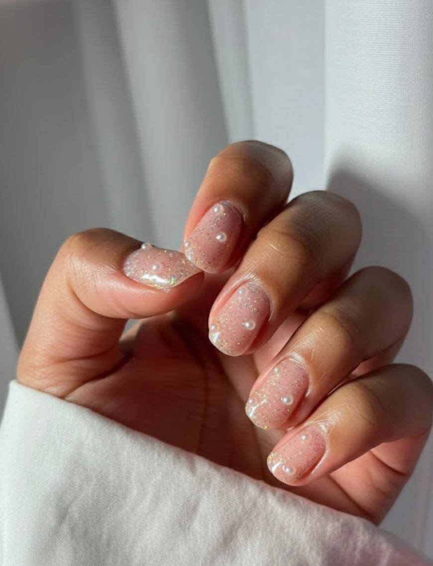 A hand with short, almond-shaped nails featuring sheer pink polish, glitter, and small pearl-like beads as decoration. The background is soft white curtains, and the hand is partially covered by a white sleeve.