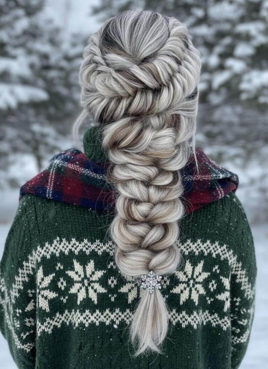 A person with long, thick, silver-blonde hair styled in an intricate braid, wearing a green sweater with white snowflake patterns and a red plaid scarf, stands outside in a snowy, forested area.