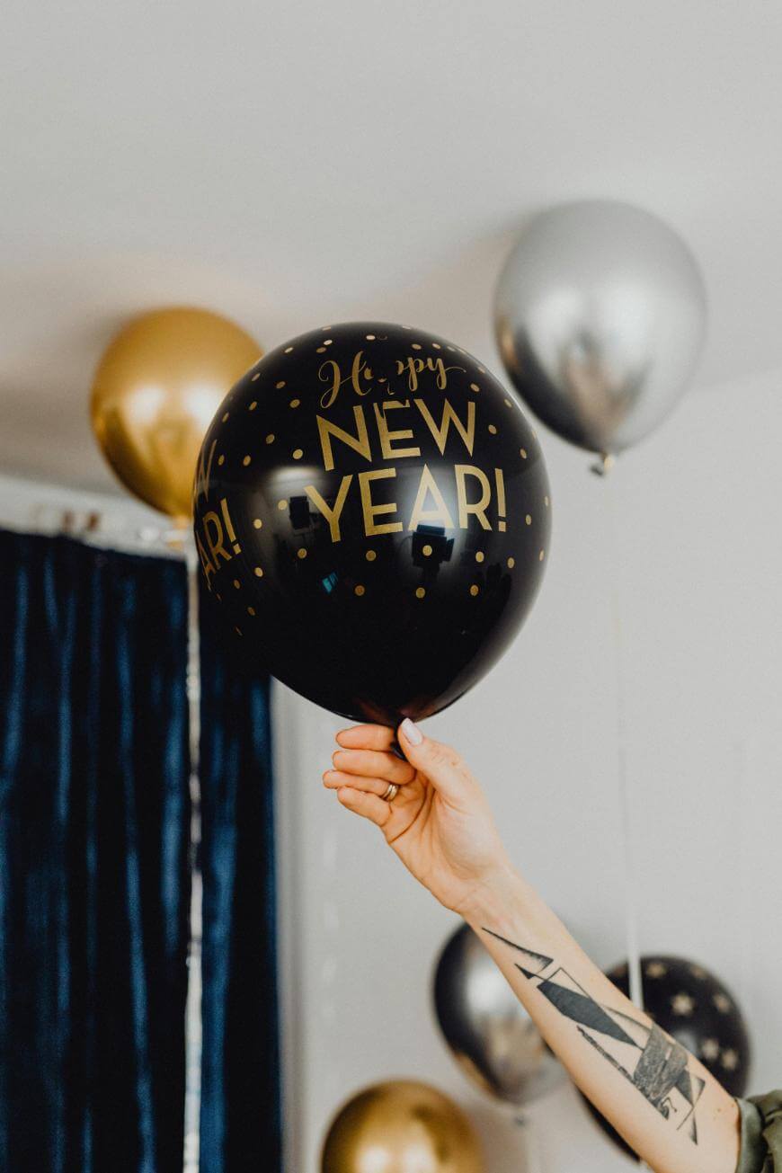 A hand with a tattoo holds up a black balloon that says Happy NEW YEAR! in gold letters, with gold and silver balloons floating in the background.