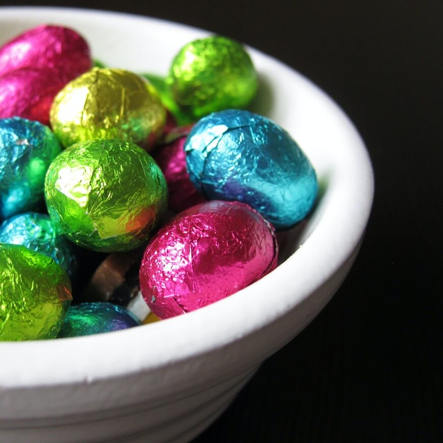 A white bowl filled with small chocolate eggs wrapped in colorful foil, including pink, blue, green, and gold, set against a dark background.