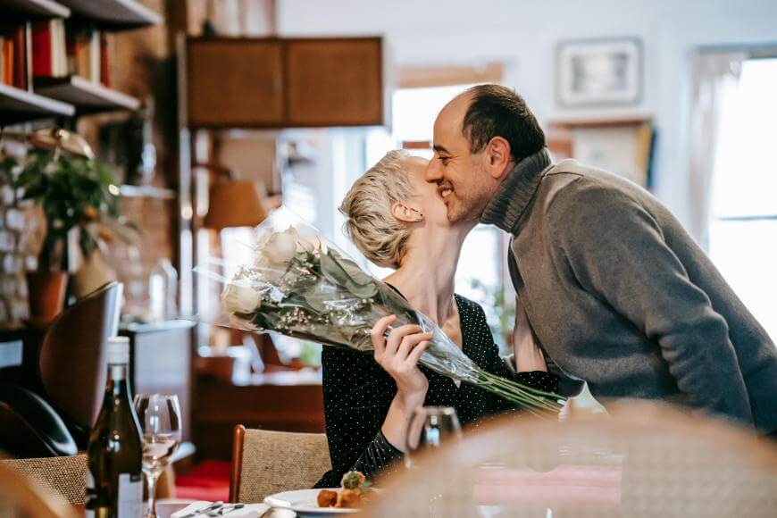 A person holding a bouquet of white roses receives a kiss on the cheek from another person, both smiling and sitting at a cozy dining table set with wine and food.