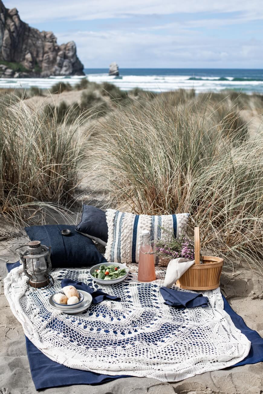 A cozy picnic setup on sandy beach dunes with a blue and white blanket, pillows, plates of food, a lantern, a wooden basket, and a bottle, with ocean waves and cliffs in the background under a blue sky.