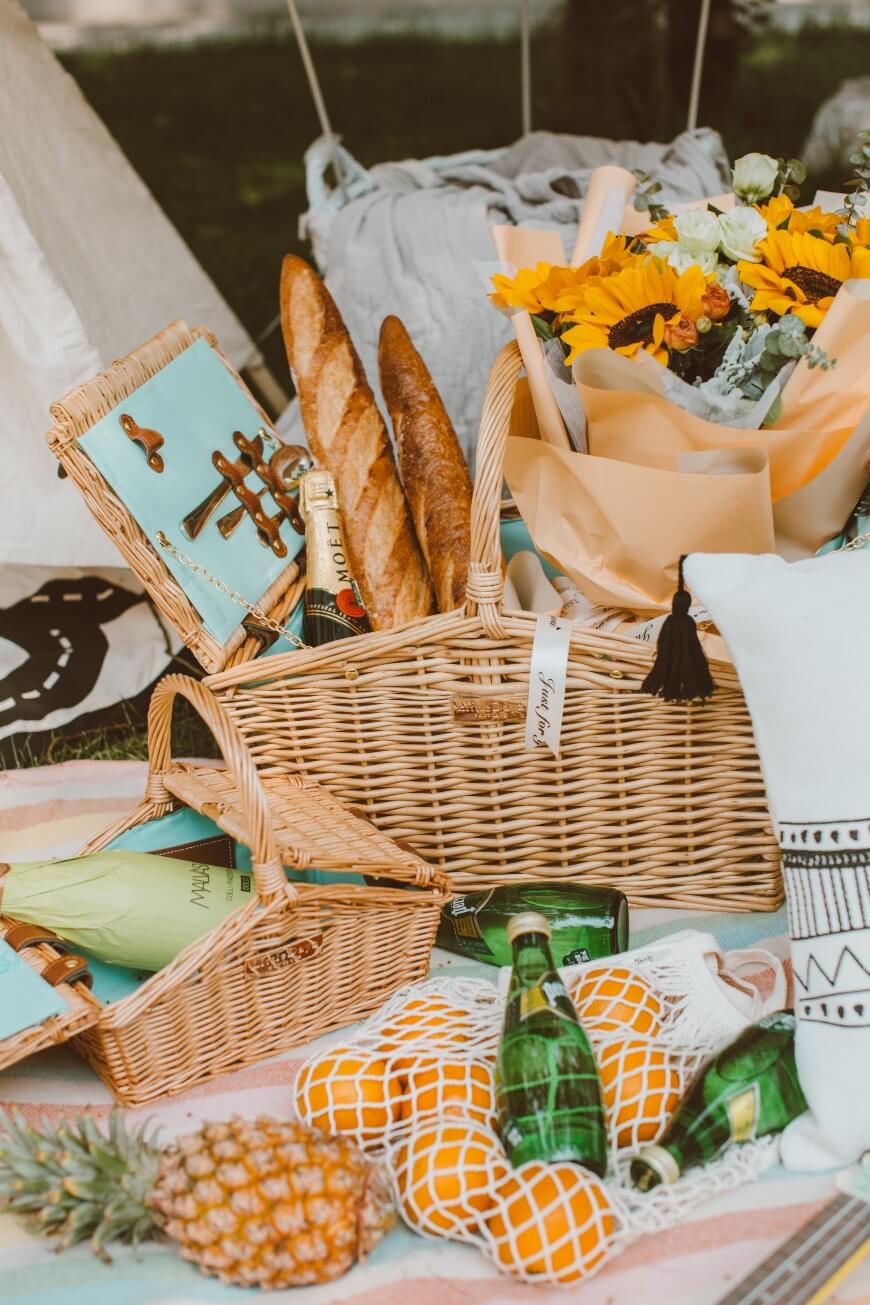 A picnic setup with wicker baskets holding baguettes, a bouquet of sunflowers, bottles, oranges in a net bag, a pineapple, and rolled-up papers, all arranged on a striped blanket outdoors.