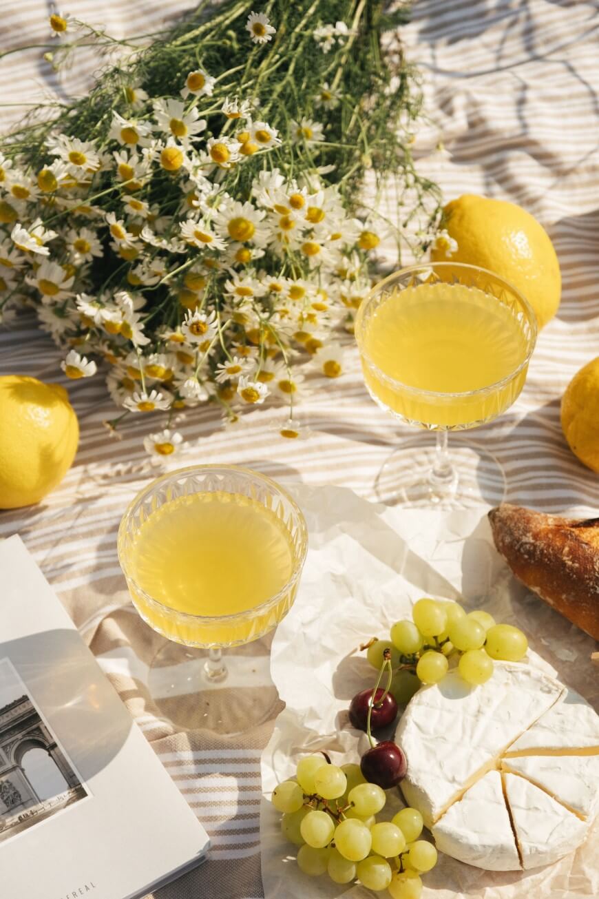 A picnic setup on a striped cloth with two glasses of yellow drink, a bunch of chamomile flowers, lemons, a baguette, green grapes, cheese, and an open book under sunlight.