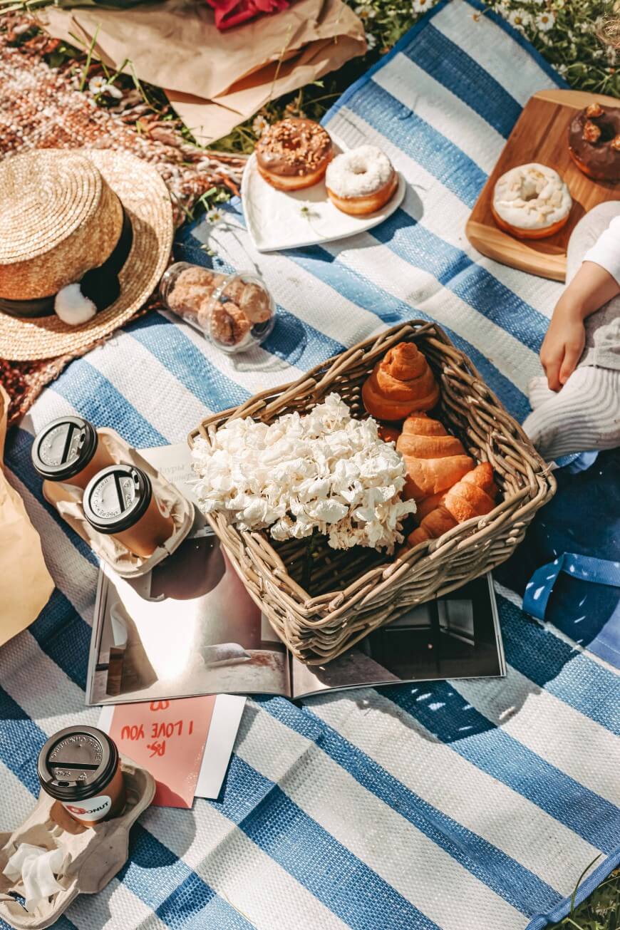 A picnic scene on a blue-and-white striped blanket with a basket of popcorn, croissants, iced coffee, donuts, a straw hat, and magazines. A person’s hand and leg are visible, reaching toward the food.