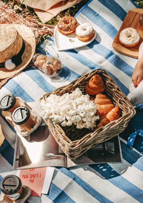 A picnic setup on a blue and white striped blanket with a basket of croissants and popcorn, donuts on plates, iced coffee drinks, a sunhat, and an open book. A childs hand reaches towards the food.