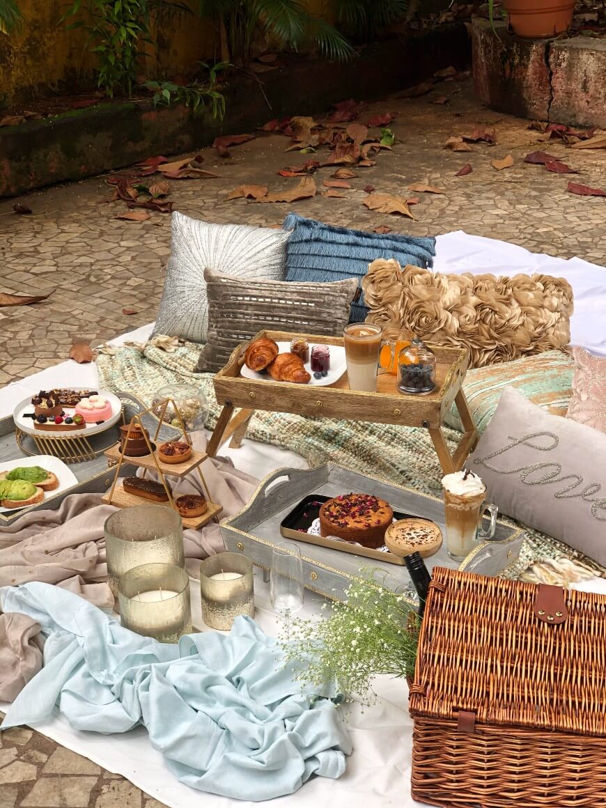 A cozy outdoor picnic setup with blankets, cushions, candles, a wicker basket, and a tray holding pastries, drinks, and desserts. Plates of food and jars with flowers are arranged on the ground. Dry leaves are scattered nearby.