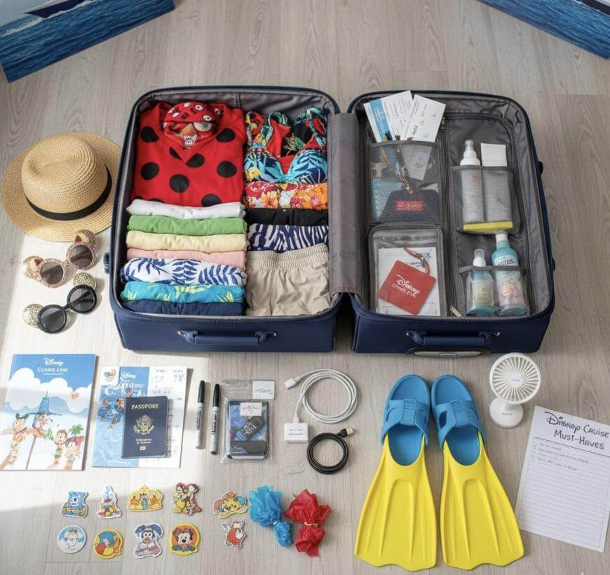 Open suitcase on floor with neatly folded clothes, travel documents, snorkel fins, hat, sandals, toiletries, gadgets, Disney cruise documents, and small accessories organized around it, ready for a Disney cruise vacation.