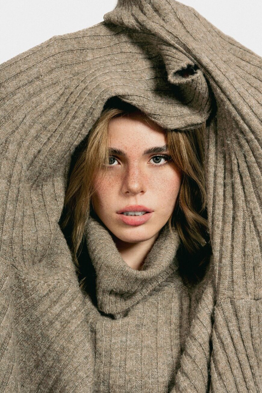 A young woman with light brown hair and freckles looks directly at the camera, framed by an oversized ribbed beige sweater draped over her head and shoulders against a white background.