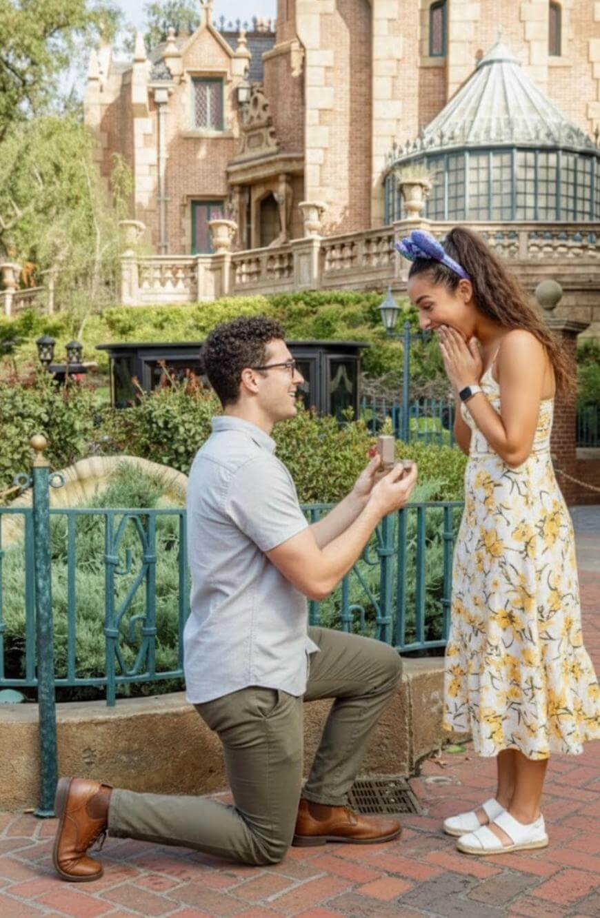 A man kneels and proposes with a ring to a surprised woman in a floral dress at a park, with a large ornate building in the background.