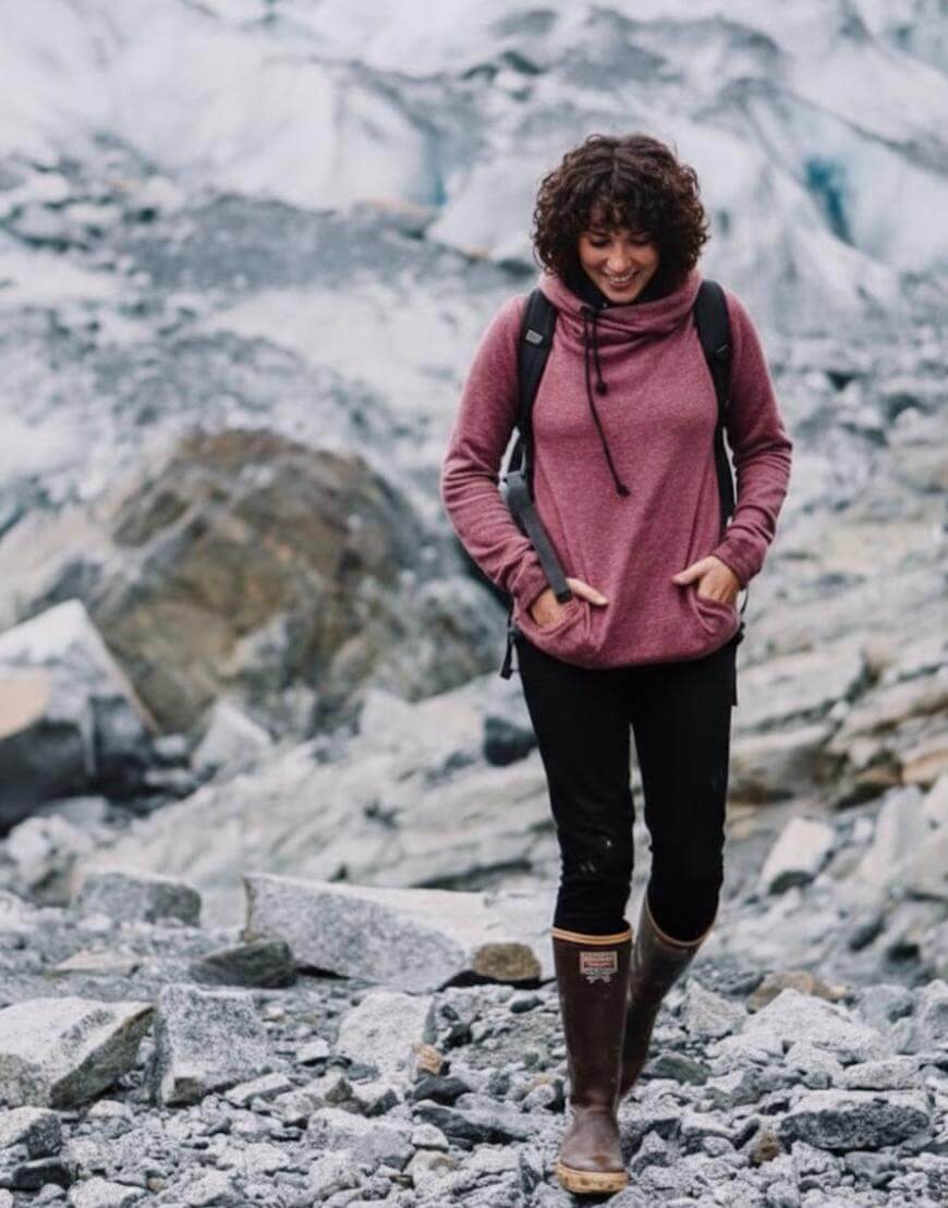 A person with curly hair, wearing a maroon hoodie, black pants, and brown boots, walks on rocky terrain with a glacier or snowy mountain in the background. They have their hands in their hoodie pockets and are smiling.
