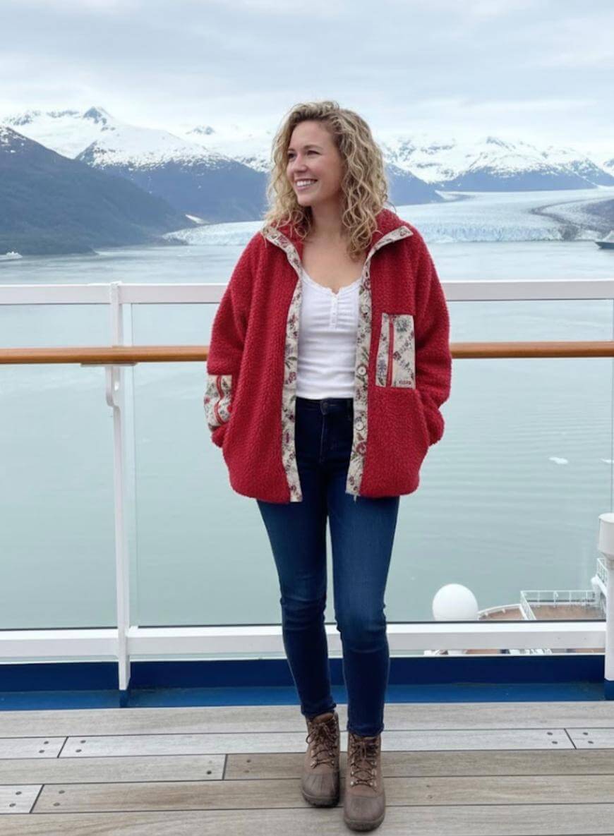 A woman in a red jacket stands on a ship deck, smiling with hands in her pockets. Snow-capped mountains and a glacier are visible across calm water in the background.