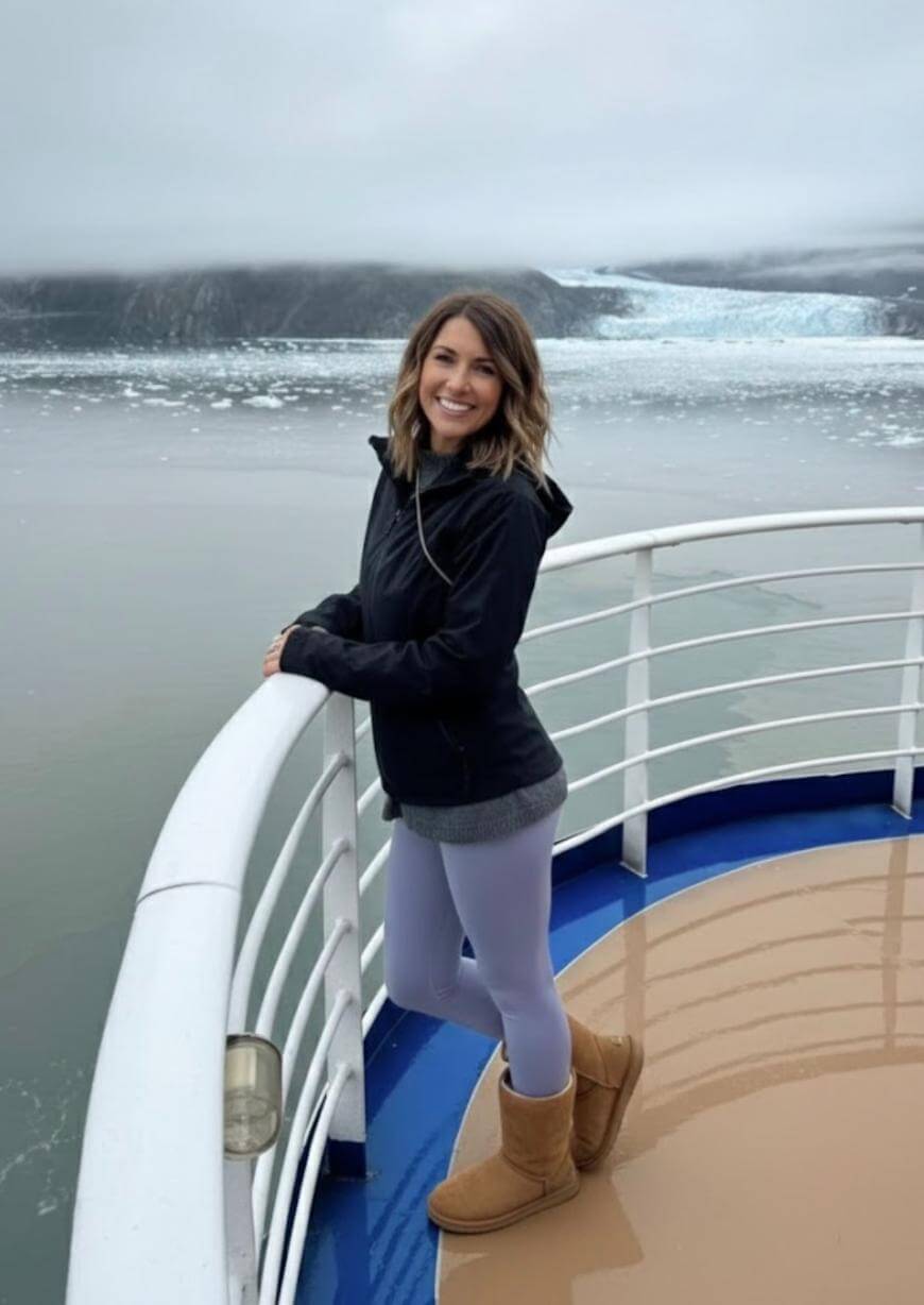 A woman in a black jacket, gray leggings, and tan boots stands smiling on a ships deck against a misty backdrop of calm water, ice, and distant glaciers.