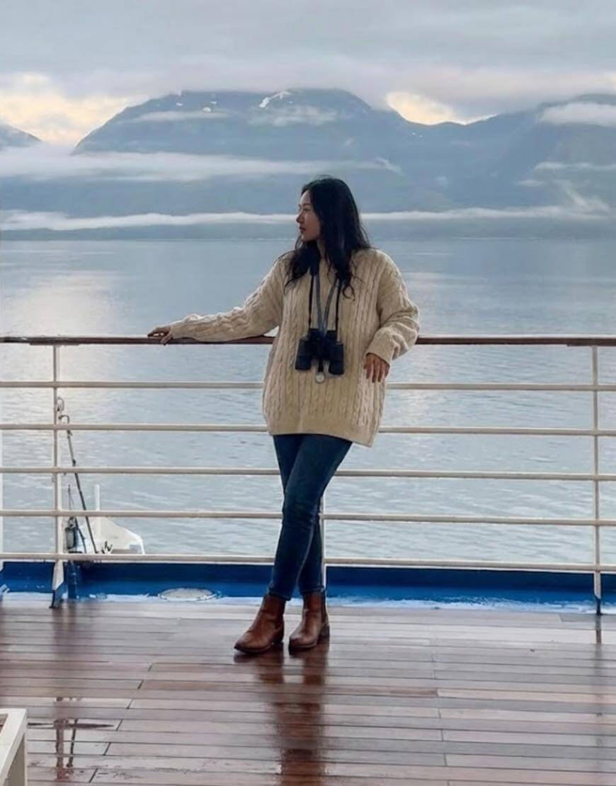 A woman in a beige sweater and jeans, with binoculars around her neck, stands on a ship deck overlooking calm water and misty mountains in the background.