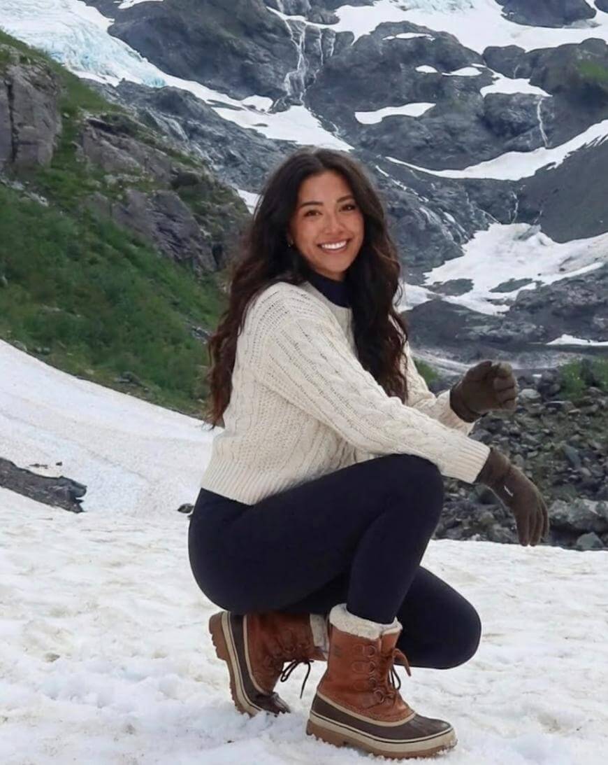A woman with long dark hair, wearing a cream sweater, black pants, gloves, and brown boots, kneels and smiles on snowy ground with a rocky, snow-covered mountain in the background.