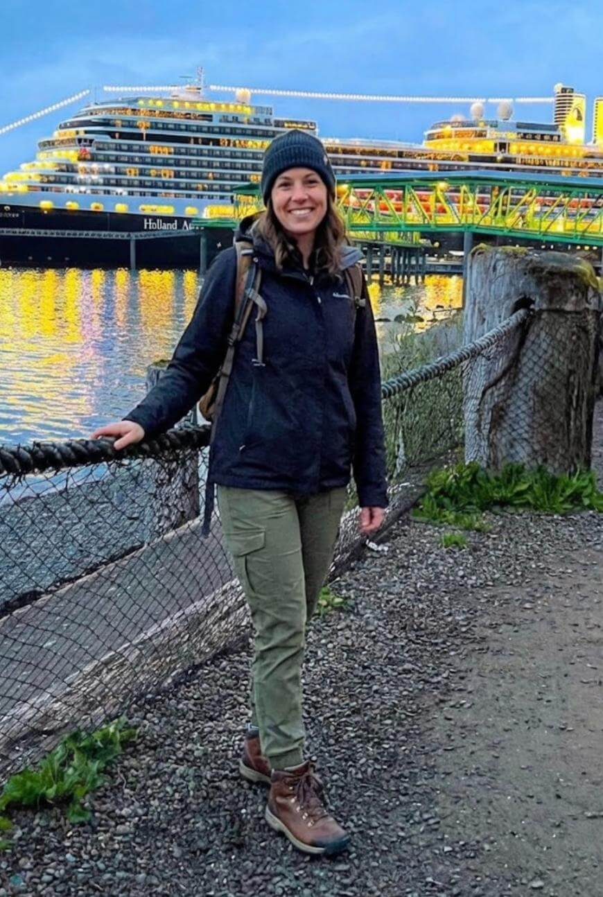 A woman in outdoor clothing and a beanie stands smiling by a fence along a waterfront, with a brightly lit cruise ship and a dock in the background at dusk.
