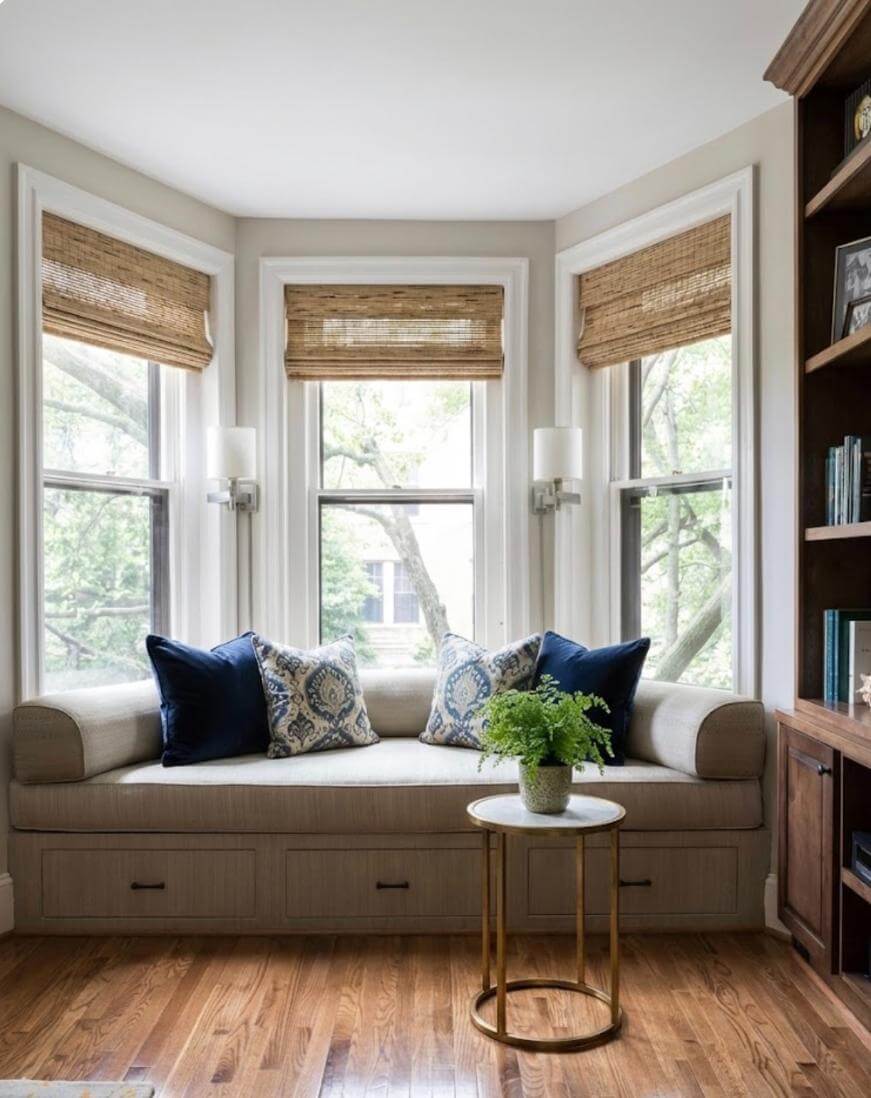 A cozy window nook with a beige cushioned bench, navy and patterned throw pillows, a round gold side table with a potted plant, and three large windows with woven shades, overlooking trees.