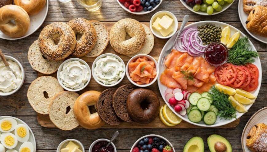 A brunch spread with assorted bagels, cream cheese, smoked salmon, sliced tomatoes, cucumbers, onions, capers, lemon, radishes, dill, avocado, boiled eggs, butter, berries, and pastries on a wooden table.